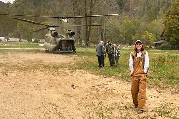 A woman in tan work overalls and a gray sweatshirt stands on a dirt road as several men converse behind her and in the distance a green Army helicopter is parked, full of supplies to be unloaded.