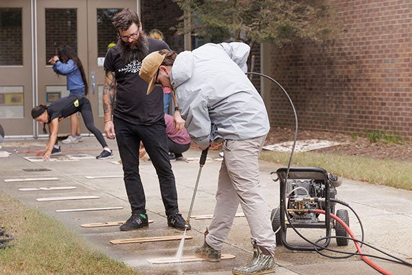 An East Carolina University student wearing a gray jacket, khaki pants and rain boots uses a power washer to clean dirt from a sidewalk around a stenciled letter as a teacher who is wearing a black shirt and jeans watches.