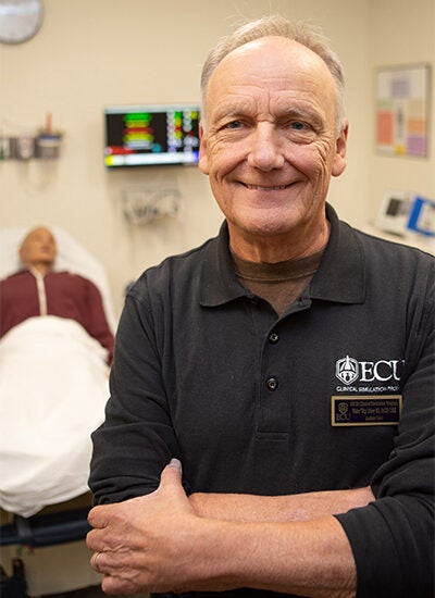 Alt Text description, no acronyms: A smiling man in a long-sleeve black shirt stands in a simulated hospital room.