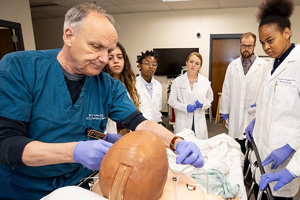 An older man in green scrubs puts clear tubing over a mannequin’s nose in a simulated hospital setting while a group of male and female students in white lab coats watch.