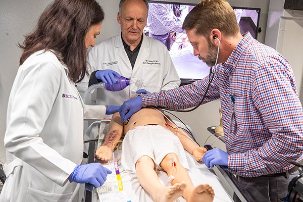 Alt Text description, no acronyms: An older man in a white lab coat holds a medical device over a mannequin’s face while a woman and man evaluate a simulated patient on a hospital bed in a simulated hospital room.