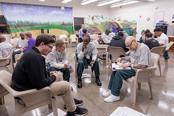  Men, the majority wearing gray East Carolina University T-shirts, sit in chairs in a small group. Some of the men are talking while another points to a paper in his lap. 