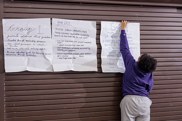 A student wearing a purple sweatshirt and gray sweatpants tapes several large pieces of paper with group notes on a wall. 