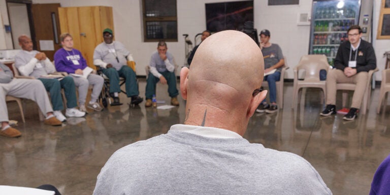 A group of men and women sit in chairs in a circle. In the center is a bald man facing the group who is wearing a gray T-shirt with black letters that spell Inside-Out Student on the back.