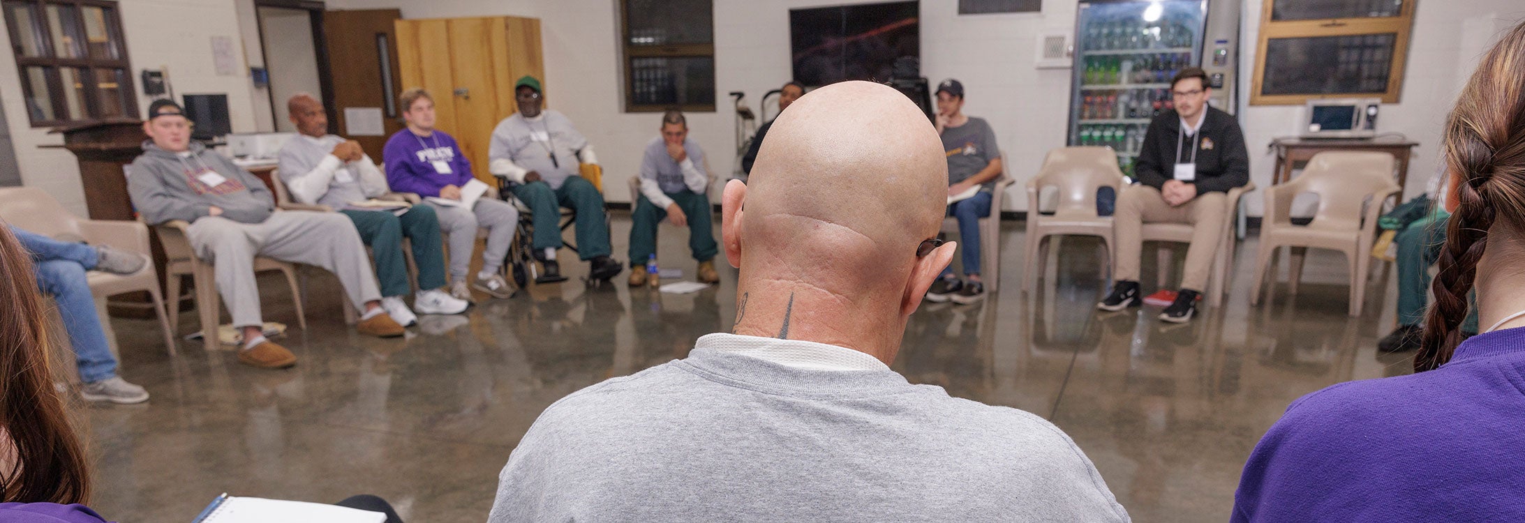 A group of men and women sit in chairs in a circle. In the center is a bald man facing the group who is wearing a gray T-shirt with black letters that spell Inside-Out Student on the back.