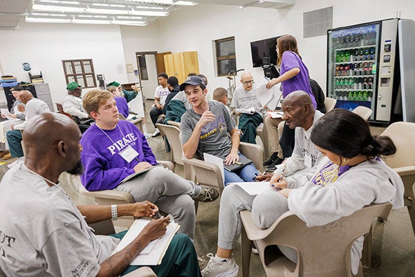  Students talk and take notes while gathered in a group of chairs in a room while other groups of students sit in separate groups.