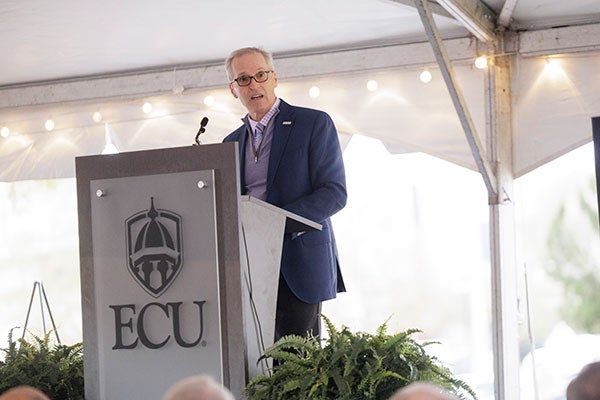 Man in sportscoat and lavender sweater vest delivers remarks at a lecturn with the emblem for East Carolina University showing.