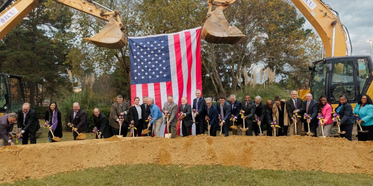 28 people stand in front of a long, low mound of sandy ceremonially shoveling dirt in front of two construction excavators strung with a large American flag.