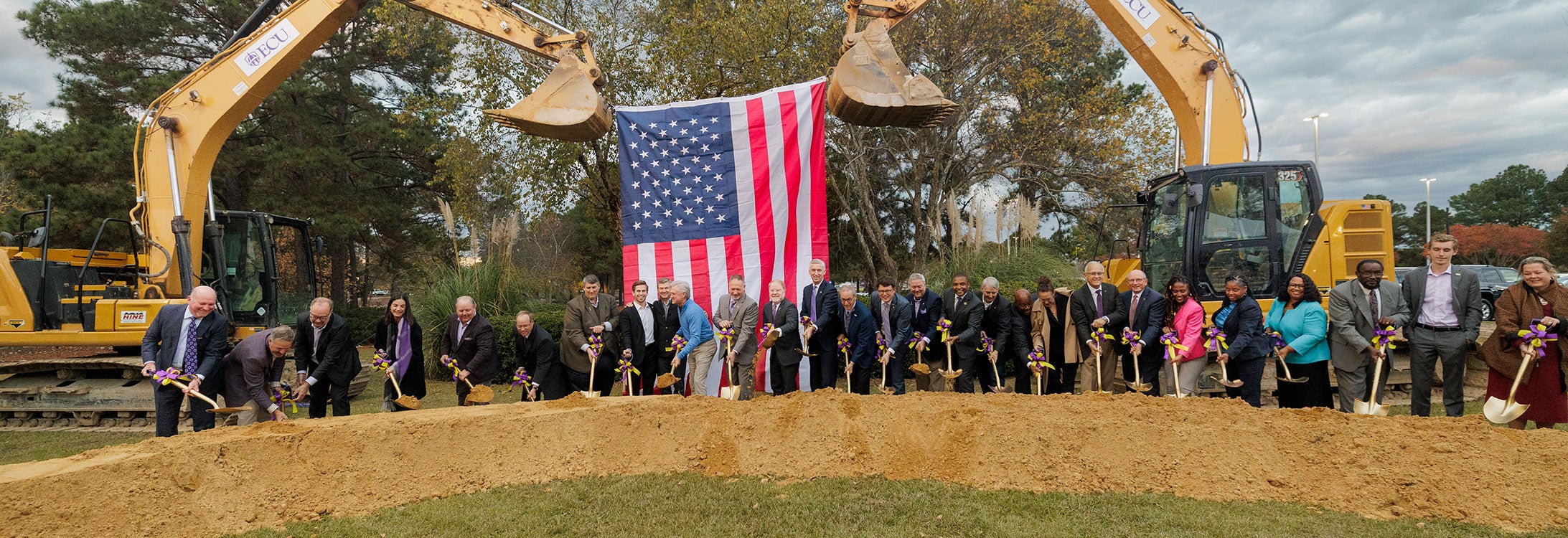 28 people stand in front of a long, low mound of sandy ceremonially shoveling dirt in front of two construction excavators strung with a large American flag.