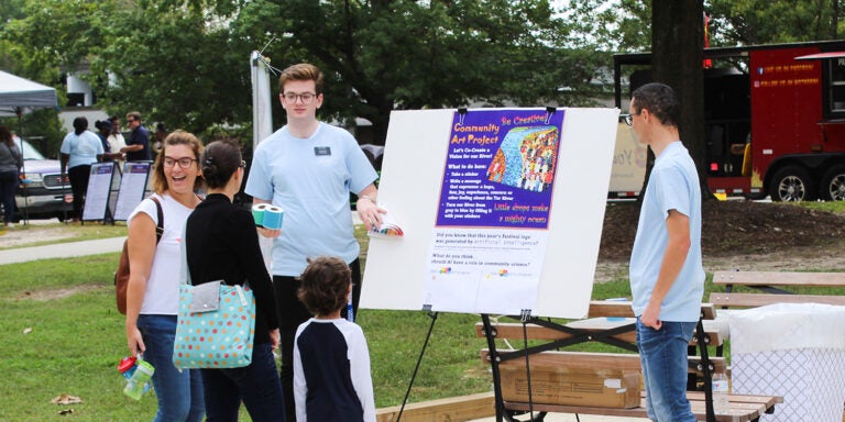 Local community families and children talk to an East Carolina University student at an exhibit outside at the Town Common in Greenville, North Carolina, during the 2023 Tar River Community Science Festival. A red food truck and tent with more staff appear in the background.