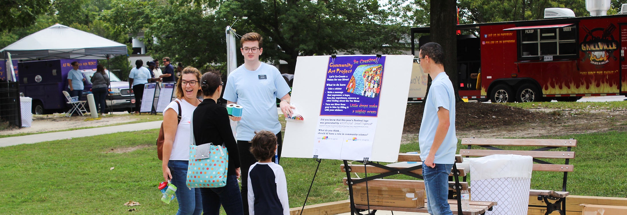 Local community families and children talk to an East Carolina University student at an exhibit outside at the Town Common in Greenville, North Carolina, during the 2023 Tar River Community Science Festival. A red food truck and tent with more staff appear in the background.