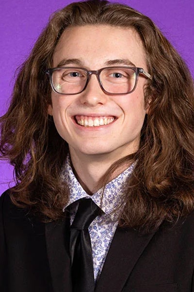 Zasha Griffin smiles in front of a purple photo backdrop. He has long, curly, brown hair, and he is wearing glasses, a light-purple flowered shirt, black tie and a black suit jacket.