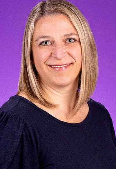 A woman with shoulder-length, straight blond hair and green eyes, is wearing a dark blue blouse and sitting in front of a purple photo background.