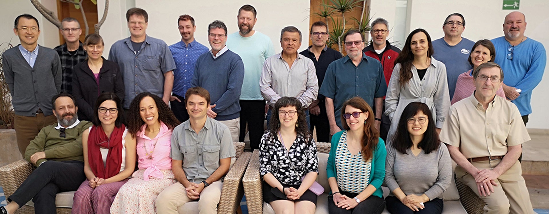 A group of nearly two dozen men and women in business casual clothes pose for a photo at the meeting of the 2024 International Council for Exploration of the Sea and the North Pacific Marine Science Organization. Some people sit on two beige wicker sofas, with the remaining people standing behind them.