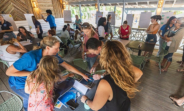 A group of scientists and community members meet under an outdoor pavilion. They are looking at charts, posters and photos on tables and spread out around the room.