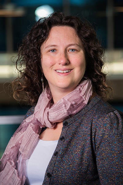 A woman with curly brown hair, is wearing a pink scarf, and a multi-colored button-up jacket over a white shirt.