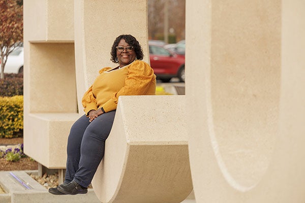 : A woman wearing a gold shirt and black pants smiles while sitting in the middle of the E-C-U letters in front of East Carolina University’s Main Campus Student Center. 