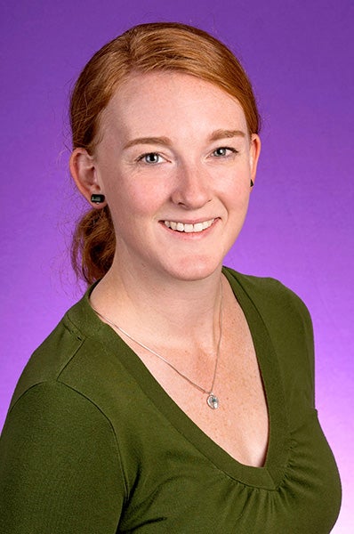 Dr. Erin Clark, wearing a green, v-neck shirt, sits in front of a purple photo backdrop.