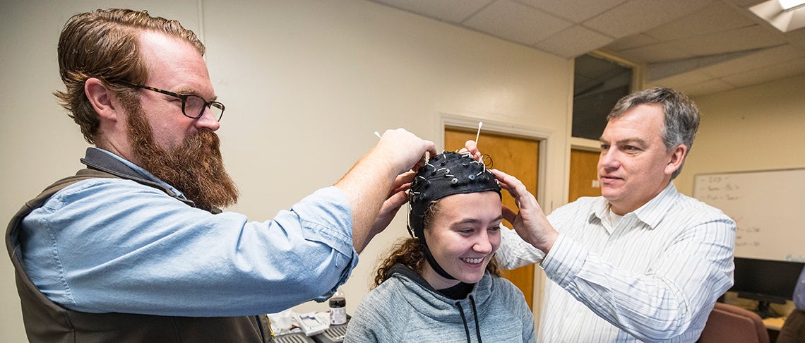 ECU faculty members Chris Mizelle, at left, and Nick Murray, at right, adjust an electroencephalography or EEG cap on ECU student Maggie Marshall’s head.