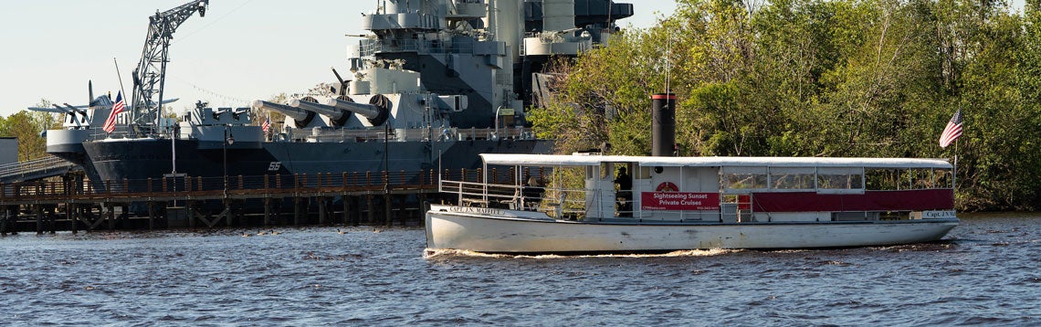 A river cruise boat passes the Battleship North Carolina