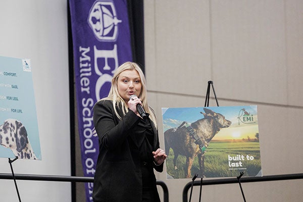 A young lady, on a stage and flanked by two posters with photos of dogs, speaks into a microphone. 