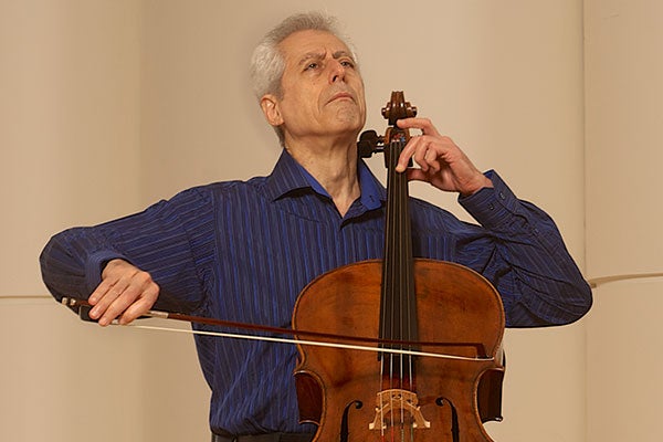 Gruber is photographed onstage with his cello in a dark blue shirt, smiling at the camera