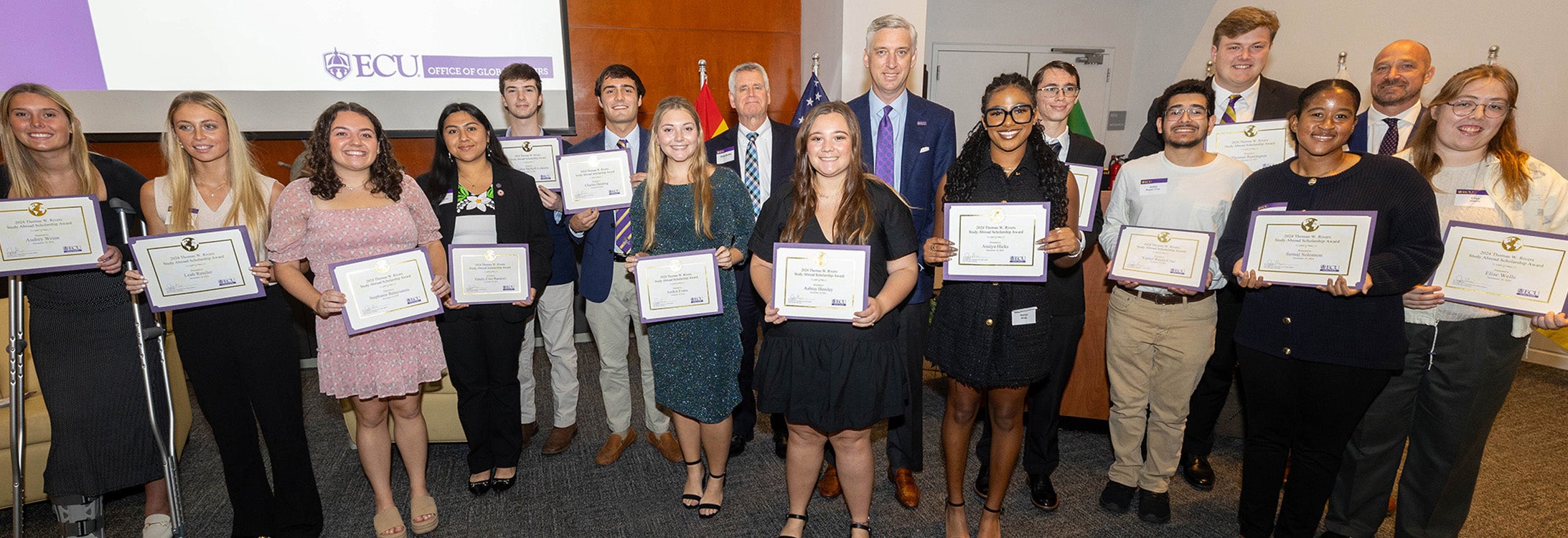 A diverse group of 15 students in business casual attire pose for a photo holding certificates with three men in suits.