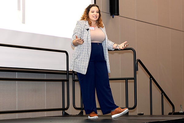 A student with brown hair explains her research on stage in front of a screen, wearing blue pants and a brown top with a plaid blazer.