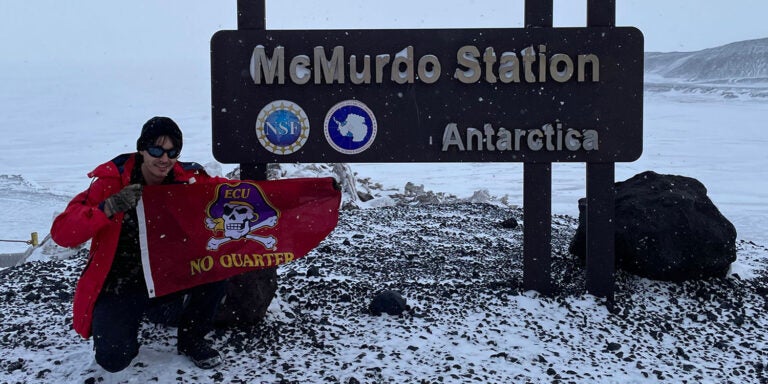 A man wearing winter outerwear holds an East Carolina University No Quarter Pirate flag in front of the McMurdo Station sign in Antarctica.