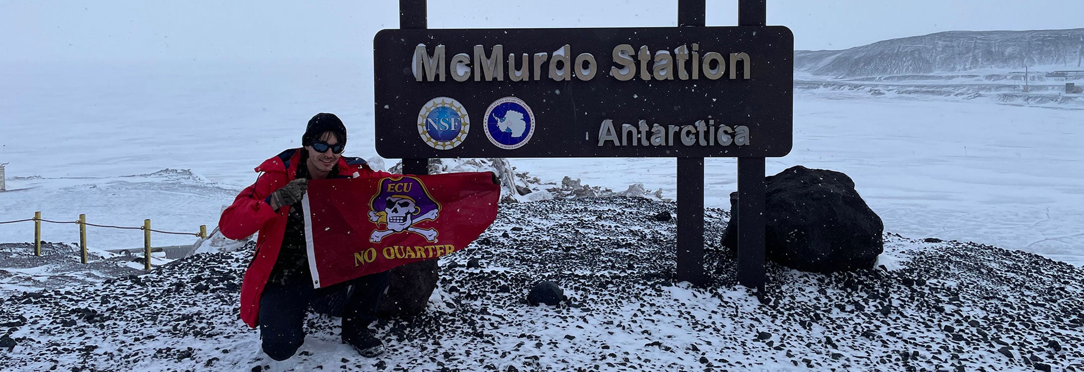 A man wearing winter outerwear holds an East Carolina University No Quarter Pirate flag in front of the McMurdo Station sign in Antarctica.