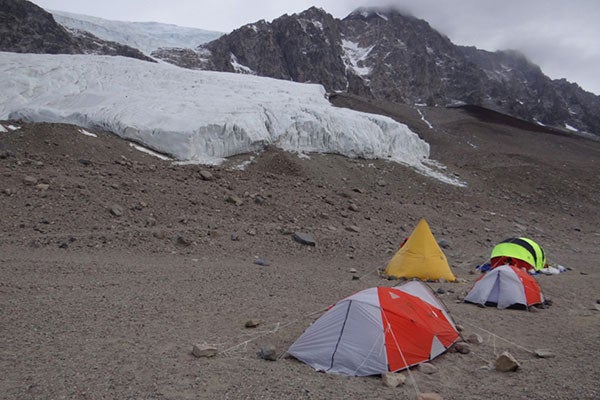 Four tents, two yellow and two white and orange, are in the foreground on rocky soil. In the background is a large glacier and mountains.