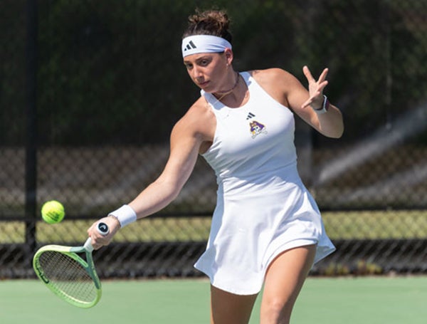 A woman in a white tennis outfit looks at the ball as she prepares to hit it over the net.