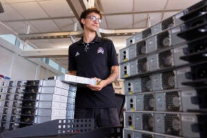 A young man wearing a black shirt lifts a computer network server from a stack of servers on a table.