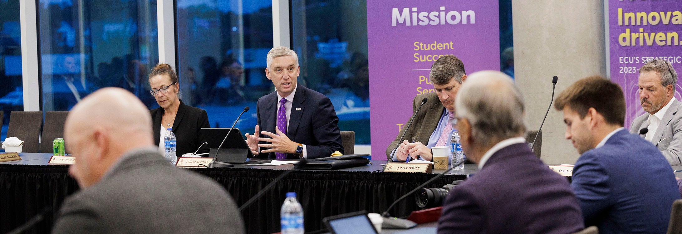 : A man wearing a dark suit is sitting at a table holding up his hands while speaking to others seated around the table.