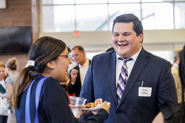 Cole Miller, a male with dark hair wears a white button up shirt, blue suit jacket, and purple and gold tie as he smiles and talks with a female student with dark hair at an event on campus.