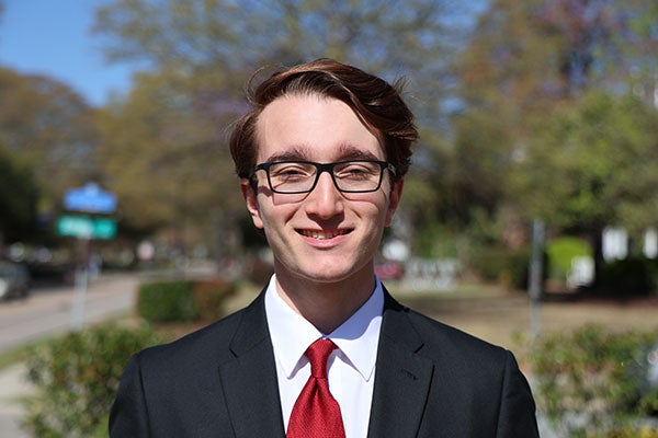 Young man in a black suit, white shirt, and red tie smiling outdoors on a sunny day, with blurred greenery and a street in the background.