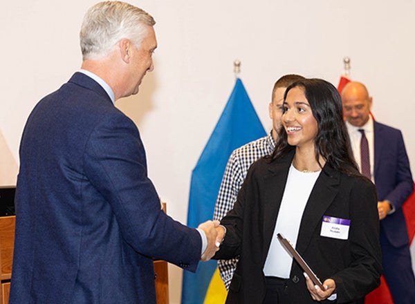 A young woman with dark hair in a black pant suit shakes the hand of a main in a suit while accepting a plaque. 