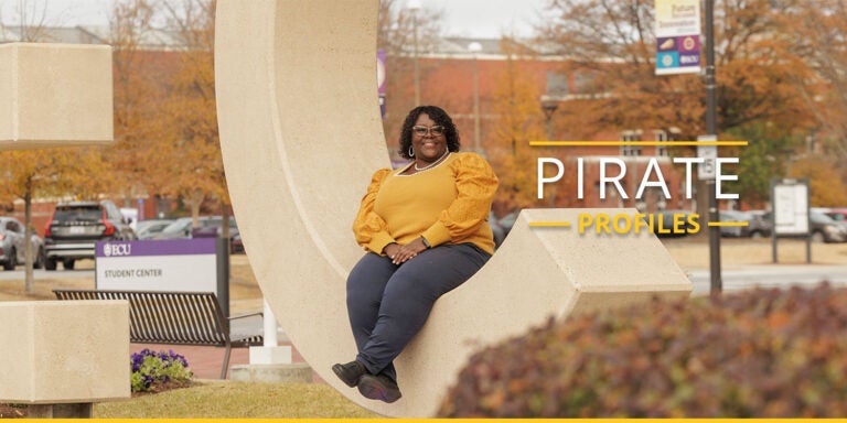 A woman wearing a gold top with black pants sits in a huge letter ‘C’ that is part of the E-C-U letters in front of East Carolina University’s Main Campus Student Center.