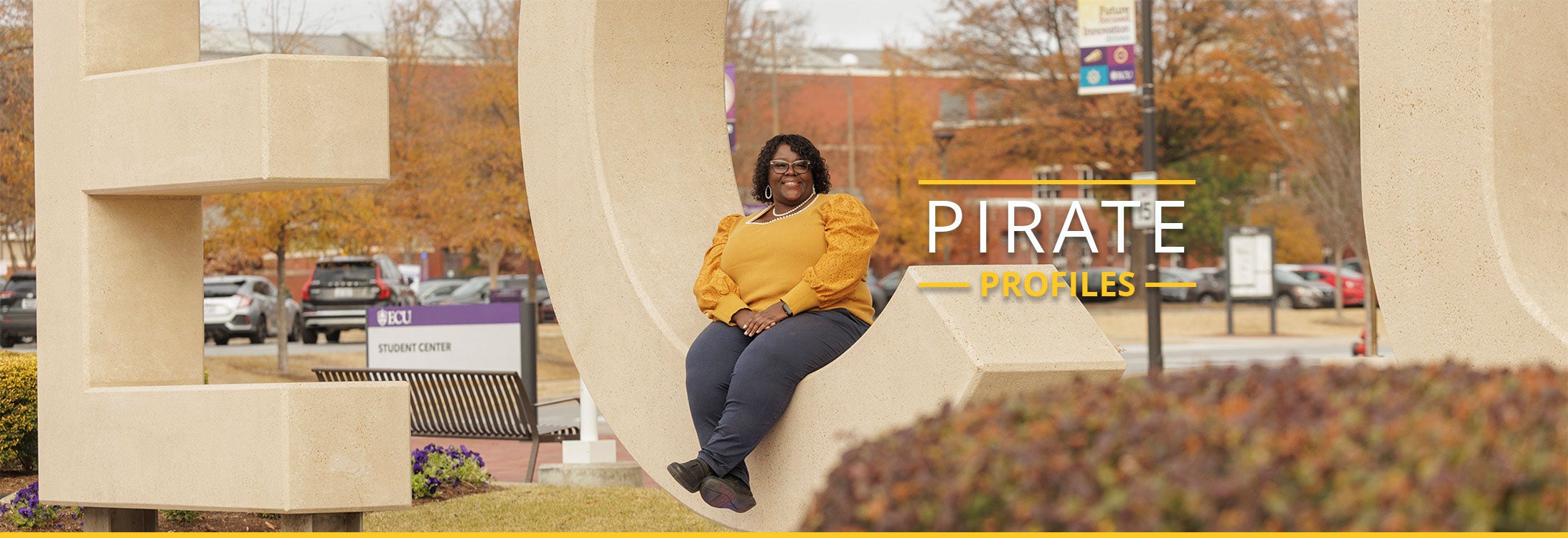 A woman wearing a gold top with black pants sits in a huge letter ‘C’ that is part of the E-C-U letters in front of East Carolina University’s Main Campus Student Center.