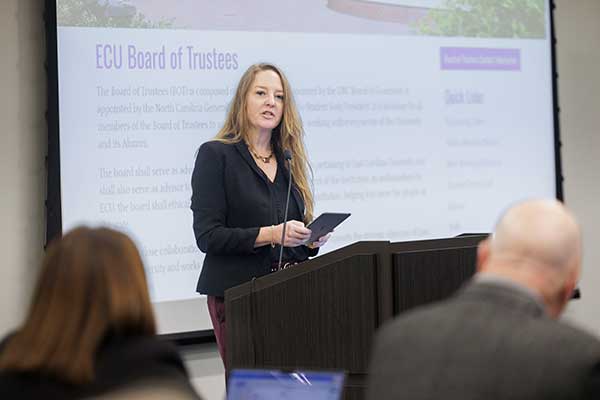 A woman in a dark jacket speaks while standing behind a podium and holds an iPad. 