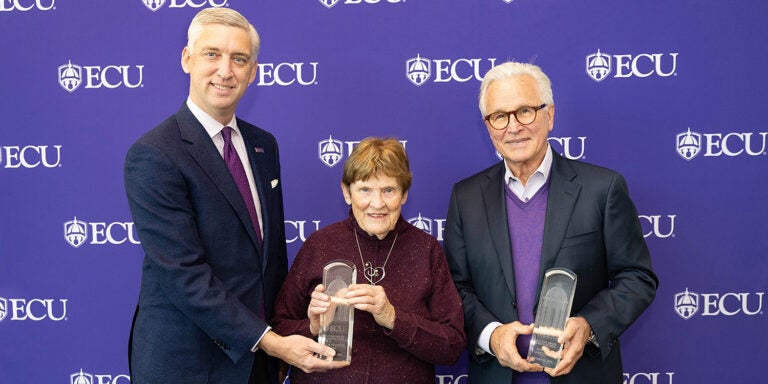 A tall man wearing a suit stands next to an older woman wearing a maroon sweater holding an award. An older man wearing a purple sweater holding an award stands to their right.