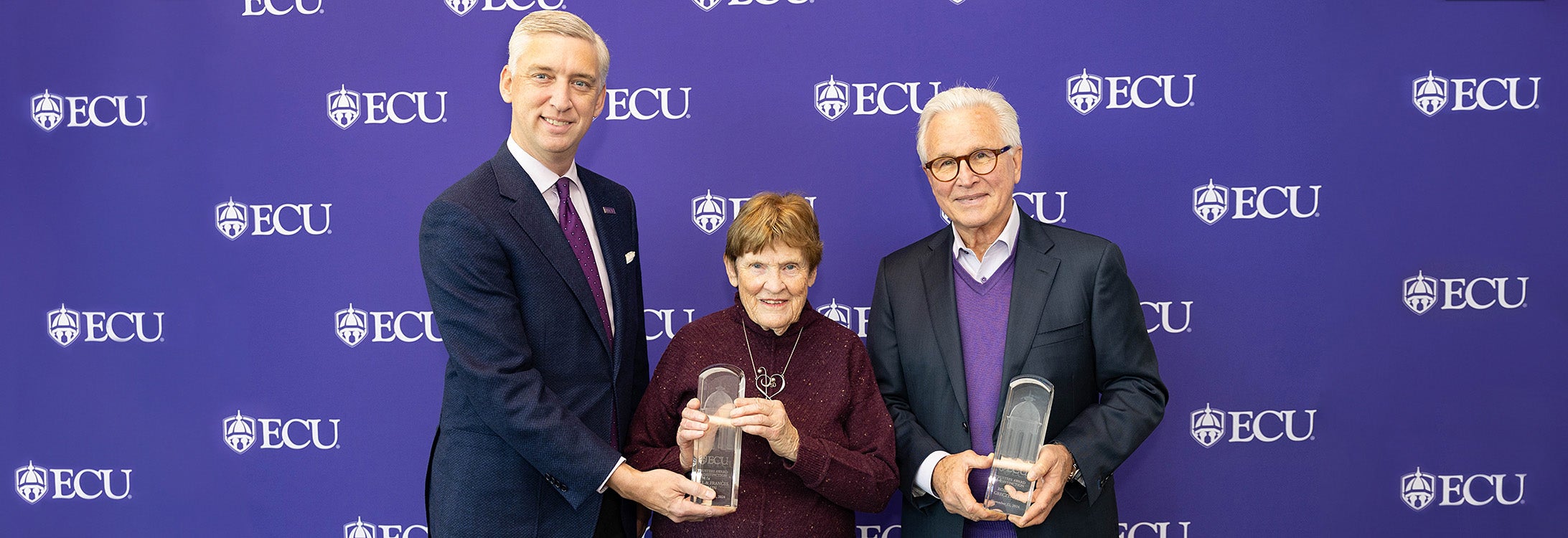 A tall man wearing a suit stands next to an older woman wearing a maroon sweater holding an award. An older man wearing a purple sweater holding an award stands to their right.