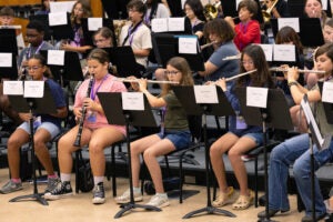 Students sit behind music stands on risers, playing woodwind and brass instruments.