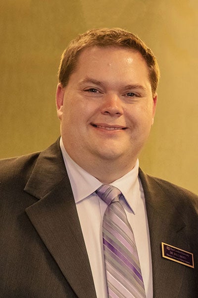 A man in a dark suit jacket and striped, purple tie smiles for a portrait photograph.