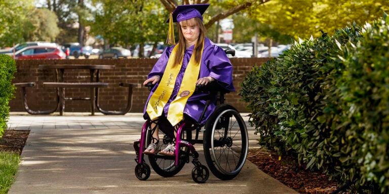 Deallaney Hudson wears purple and gold graduation cap and gown in outdoor setting beside bushes.