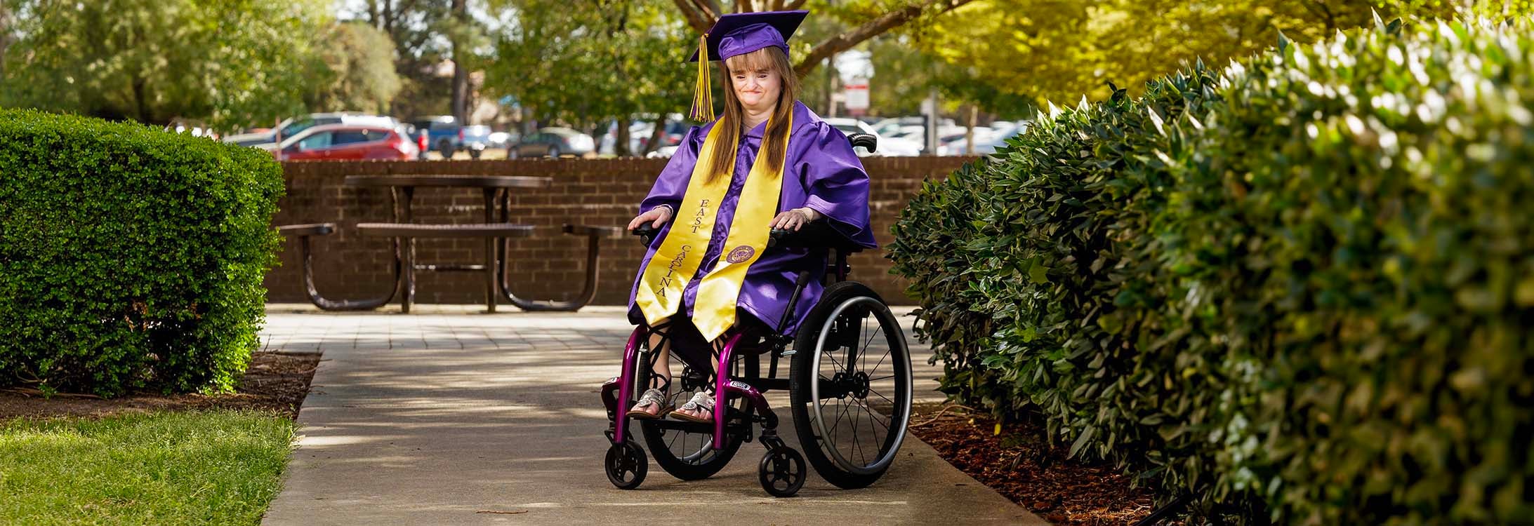 Deallaney Hudson wears purple and gold graduation cap and gown in outdoor setting beside bushes.