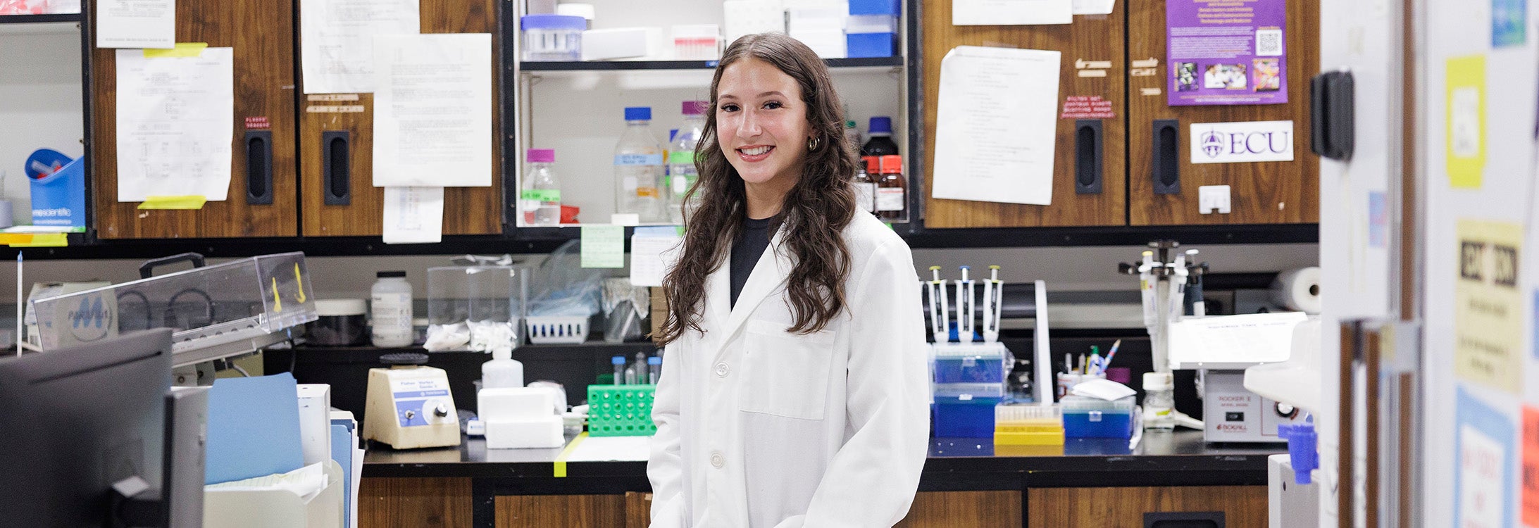 Bayli Locklear, wearing a white lab coat, stands in front of a countertop full of scientific equipment.