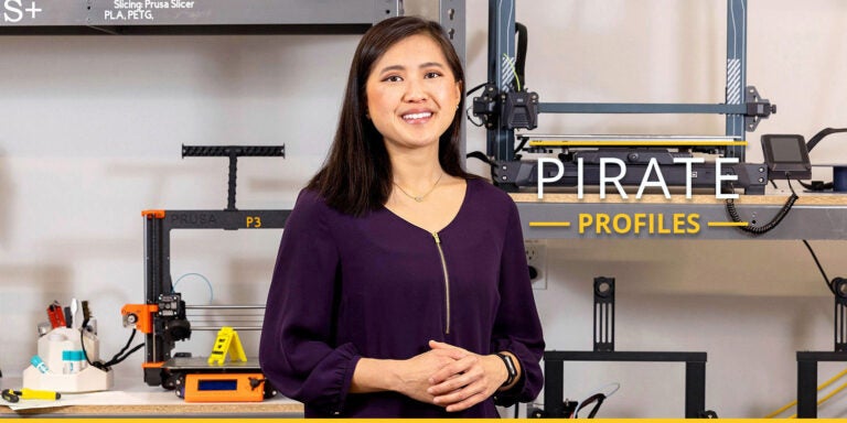 A smiling student with straight, brown hair poses in front of shelves displaying orange and black 3D printers.