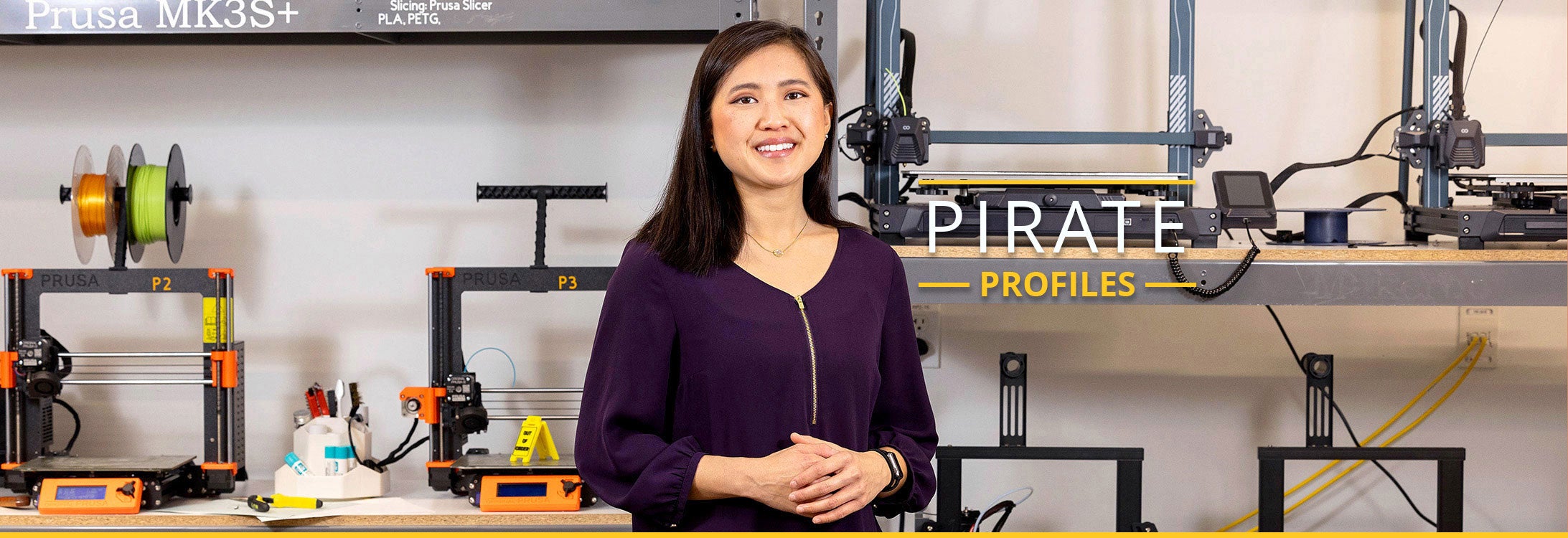 A smiling student with straight, brown hair poses in front of shelves displaying orange and black 3D printers.