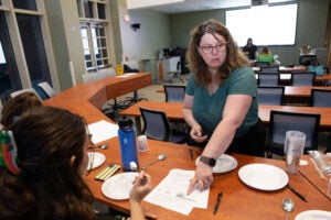 A woman in a green shirt points to a piece of paper while speaking to several women in a large room.